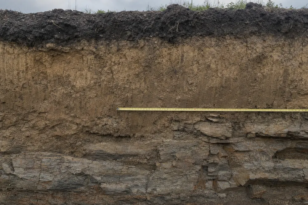 An excavation face with dark topsoil over lighter soil over layered rock, with a tape measure across it — underpinning, Sydney. Illustrative example.