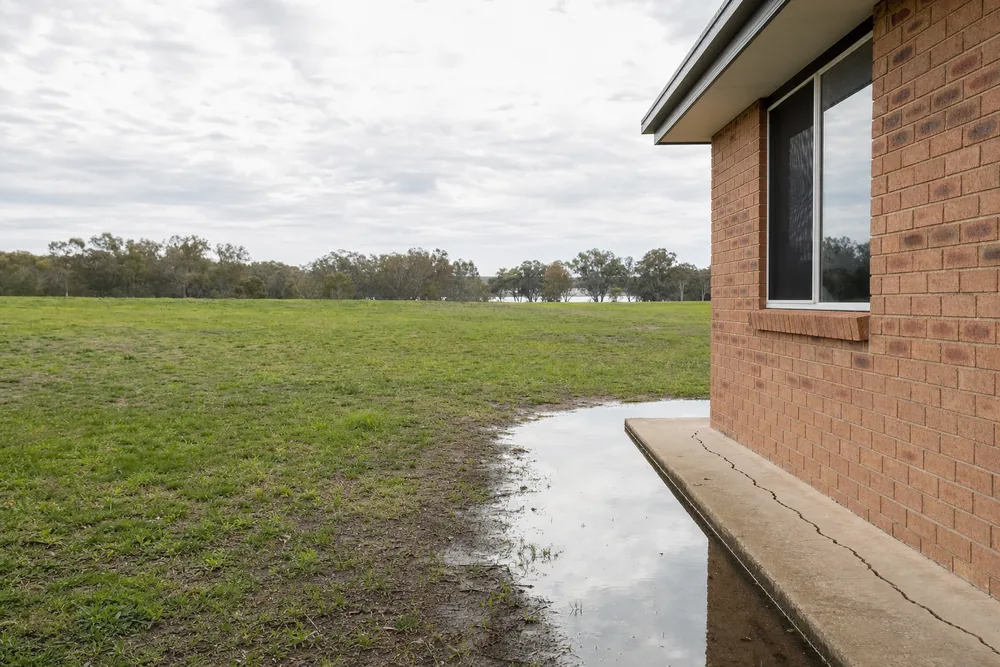 A brick house on flat ground with shallow standing water along a cracked slab edge and trees in the distance — underpinning, Sydney. Illustrative example.