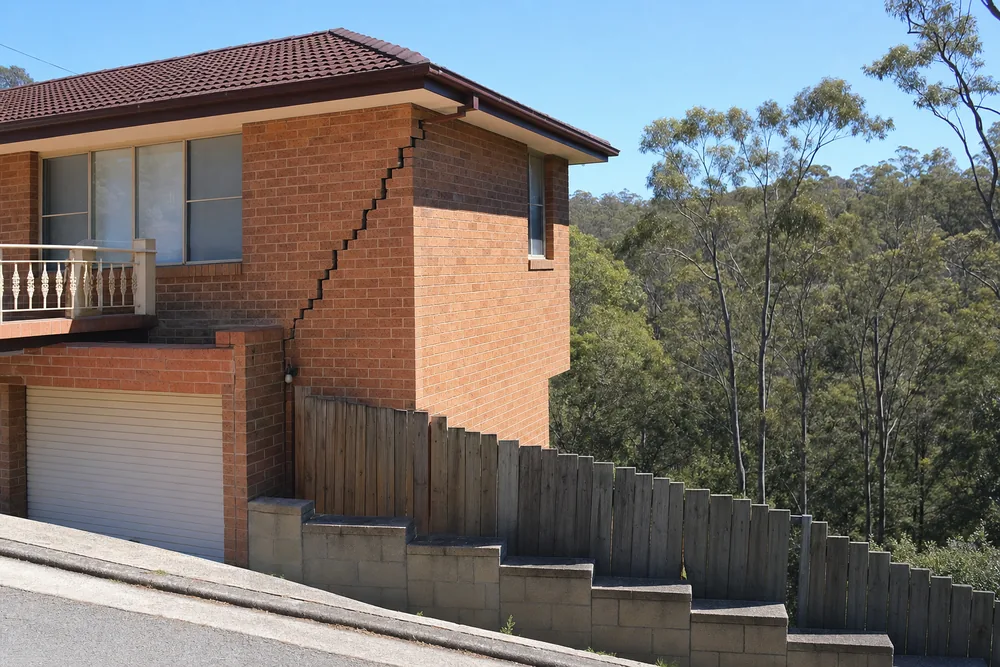 A brick house on a sloping street with a stair-step crack at its corner above a stepped block terrace — underpinning, Sydney. Illustrative example.