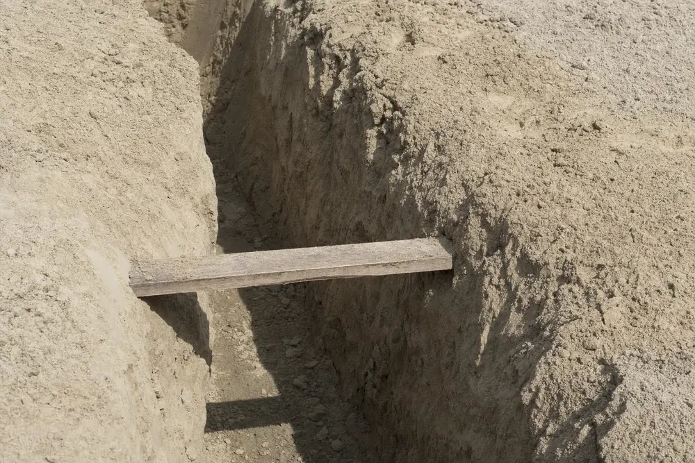 A narrow trench cut into pale, dry soil with a single plank across it — foundation repair, Sydney. Illustrative example.
